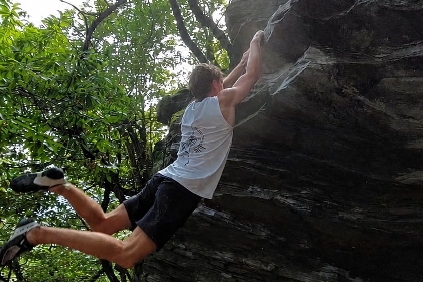 Grimpeur suspendu aux deux mains à la lèvre d'un bloc en extérieur, les pieds décollés du rocher après un mouvement dynamique