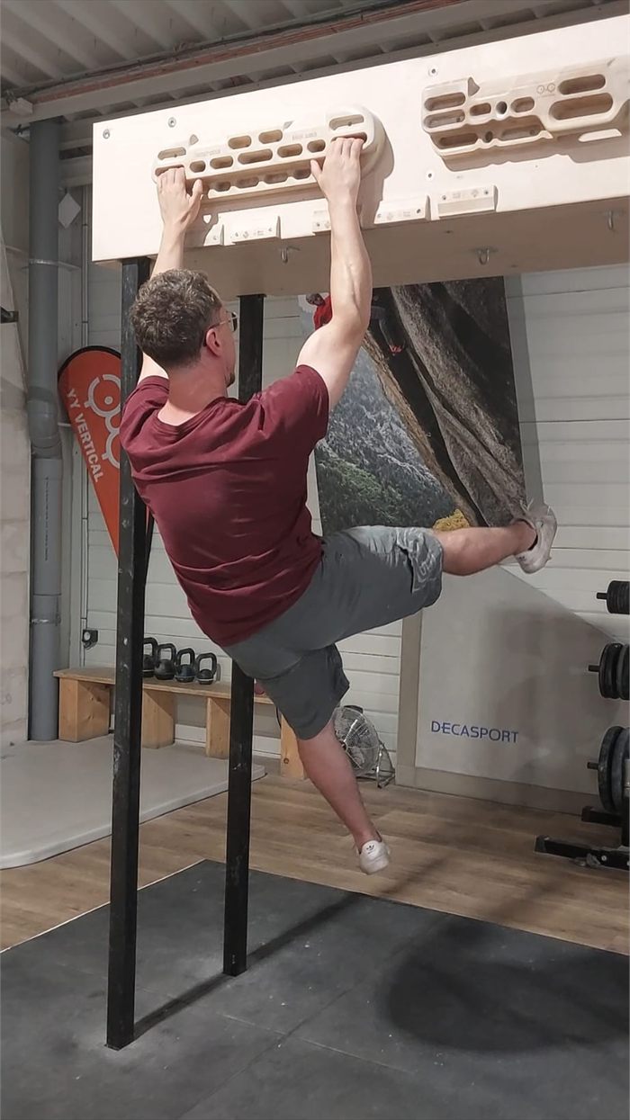 Grimpeur suspendu à une poutre, épaules fixées et omoplates serrées : la jambe monte haut et loin