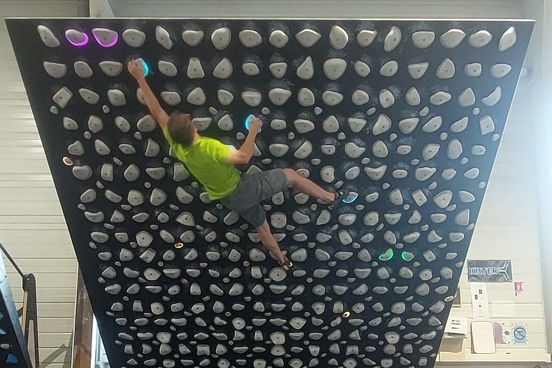 Grimpeur sur un mur très déversant en salle, un pied décollé du mur, le corps en tension entre les mains et le pied resté au contact